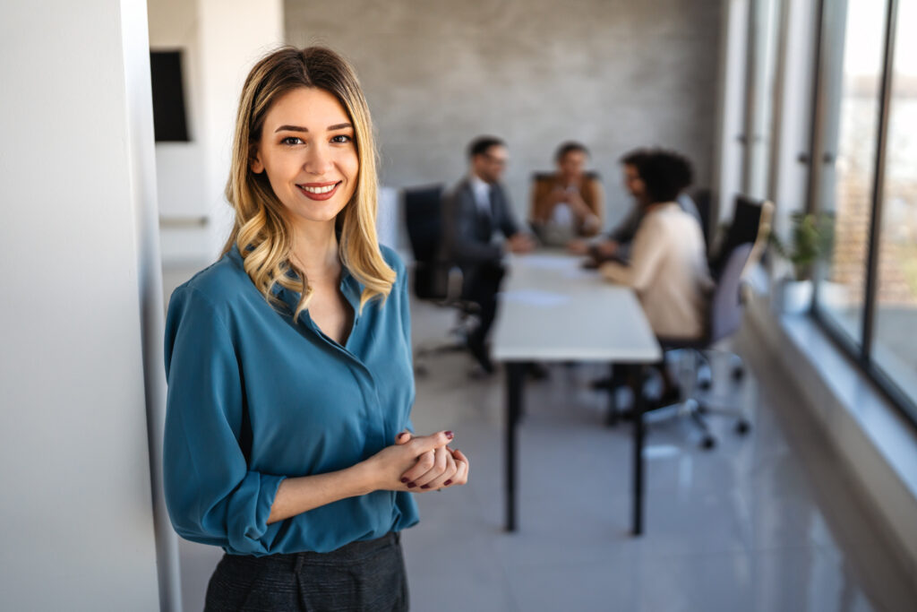 A smiling young professional standing in a modern office setting with her colleagues having a meeting in the background